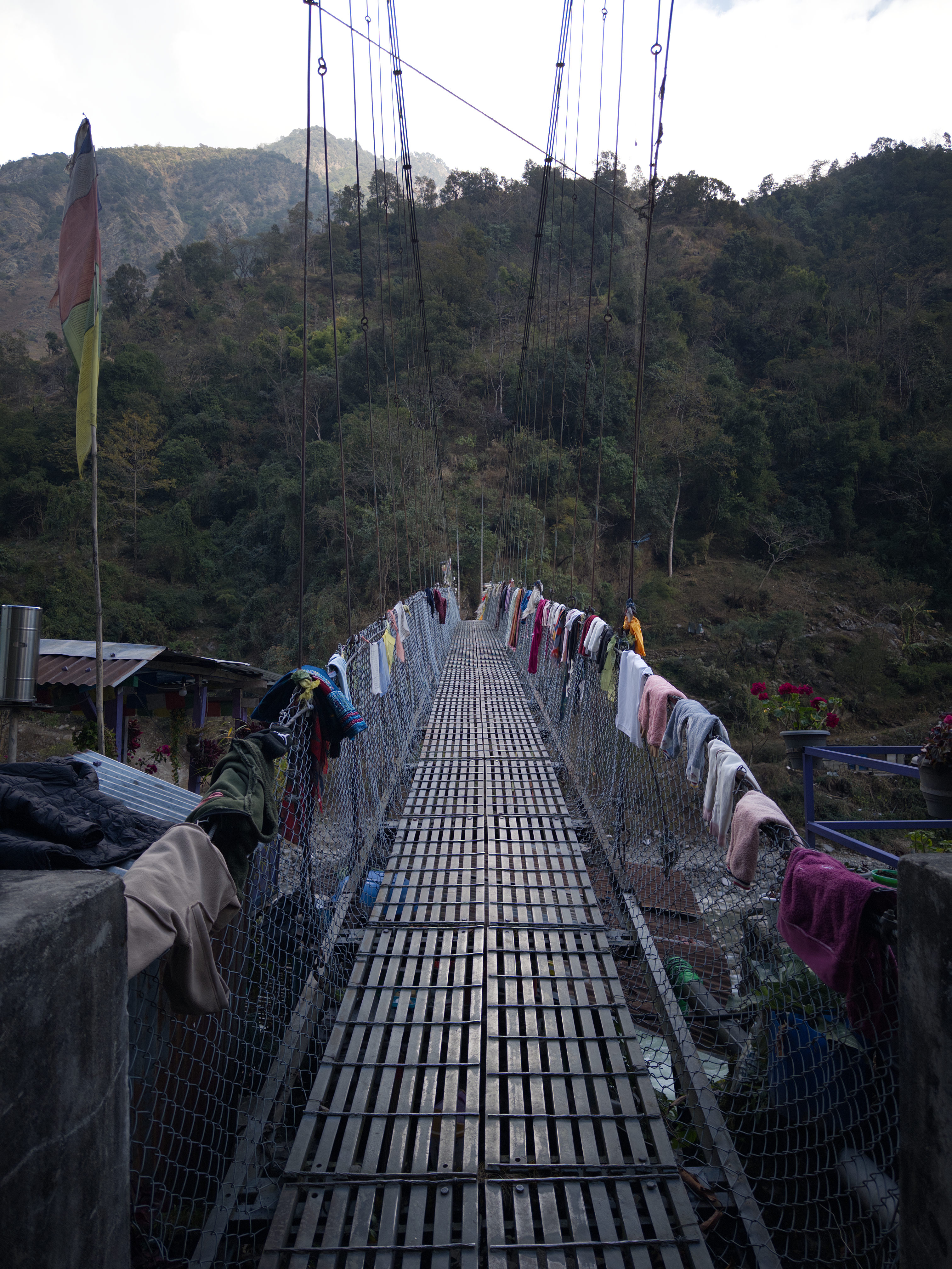 A suspension bridge stretches across a lush mountainous landscape, with clothes hanging along the railing.