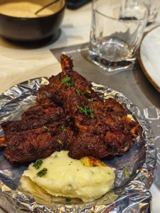 A plated dish of beef ribs on bone, garnished with fresh green herbs and served on aluminum foil.