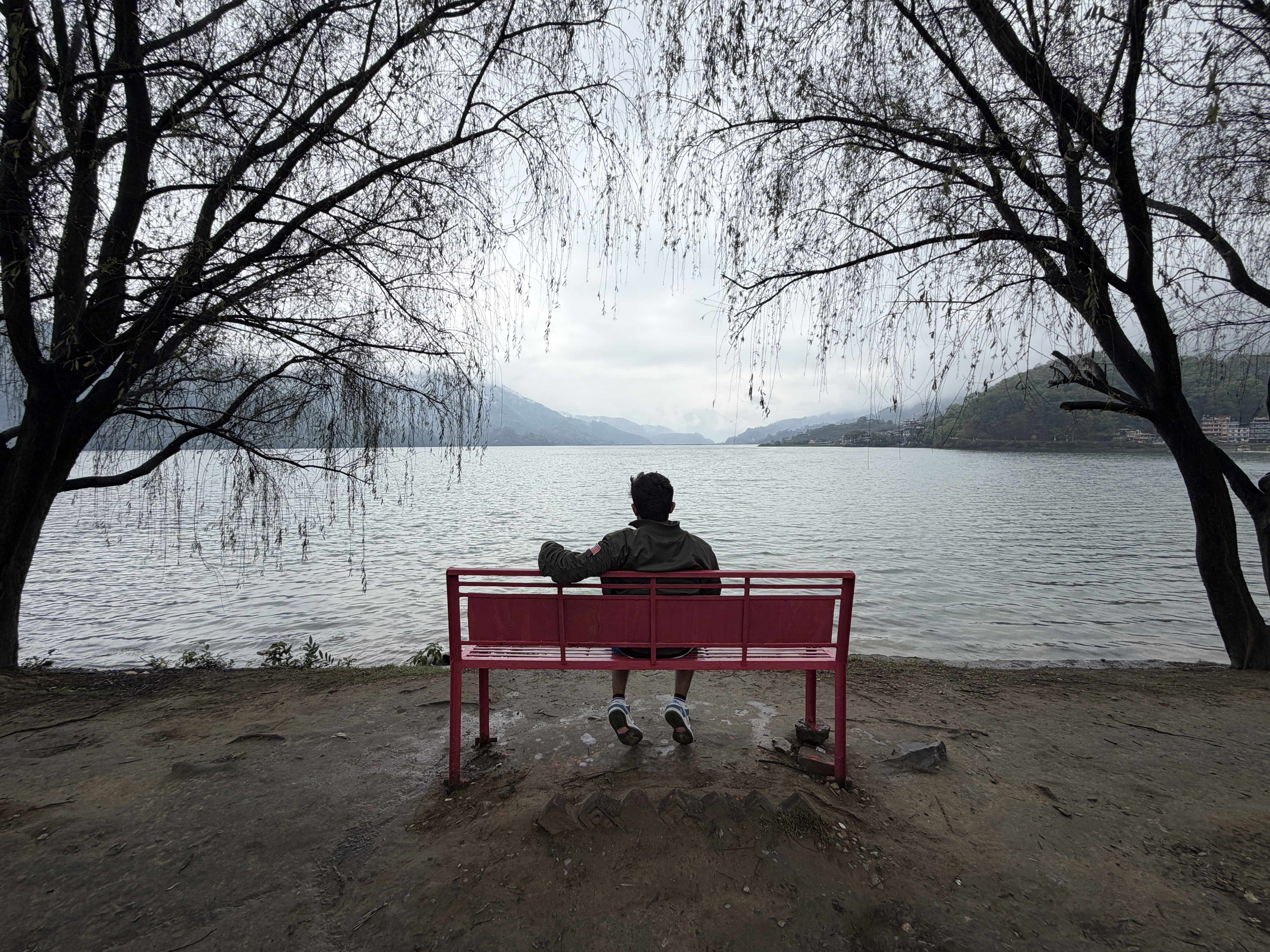 A person sits on a red bench overlooking a serene tranquil lake, framed by leafless trees. The overcast sky and distant mountains convey a calm, introspective mood.