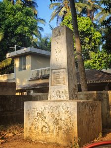 
A weathered stone monument stands tall amidst lush greenery, with palm trees and bushes nearby.