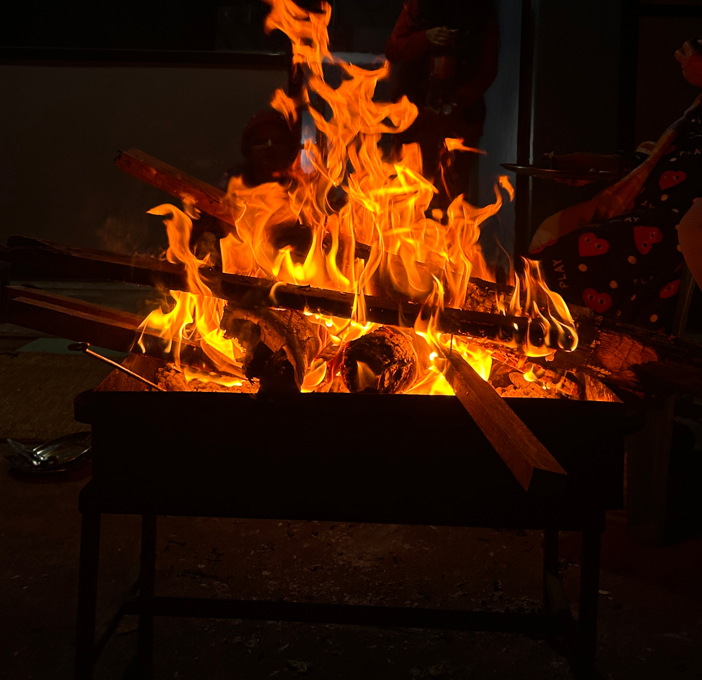 A crackling fire with bright orange flames burns atop wooden logs in a grill, creating a warm and cozy ambiance. People stand around in the shadows.