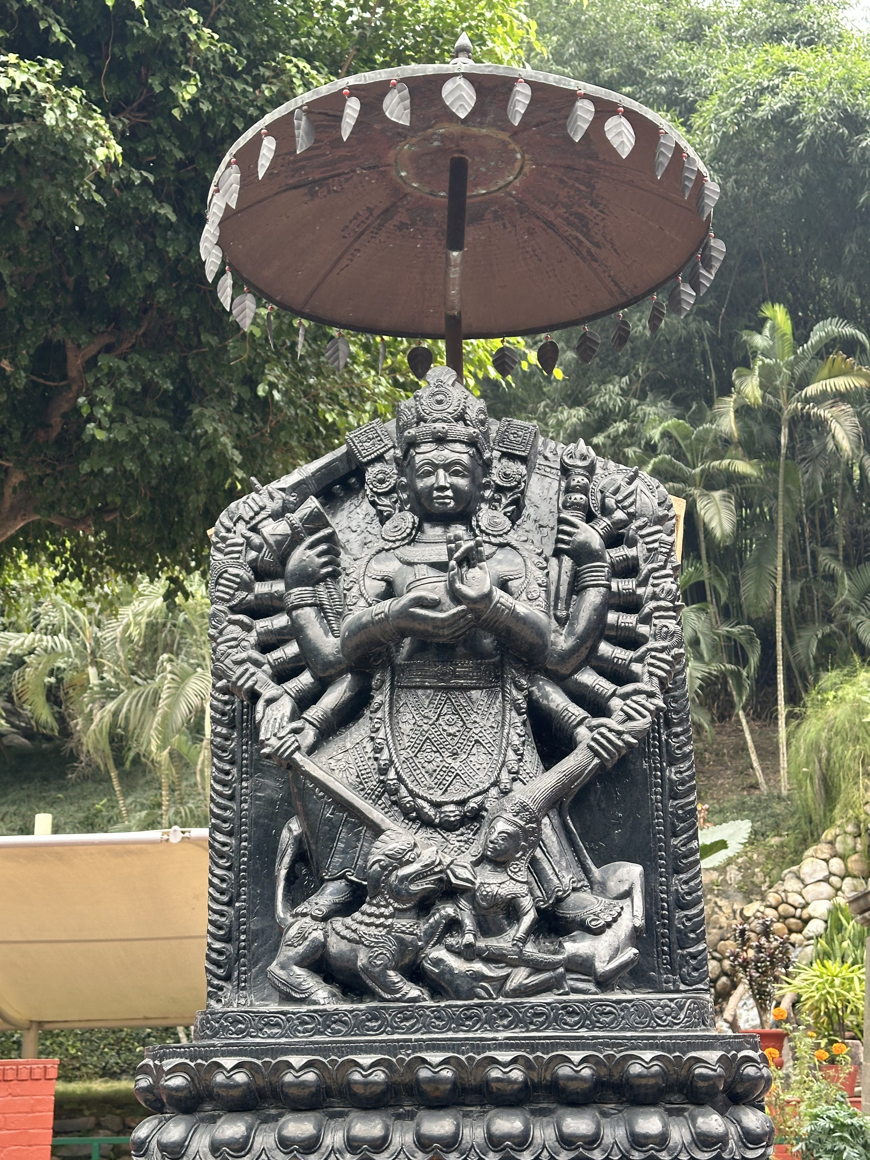 Ornate black stone statue of a multi-armed deity sheltered by a parasol, set against lush greenery.