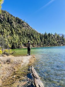 A person standing at a lake edge with arms outstretched, surrounded by lush pine trees under a clear blue sky, conveying freedom and tranquility.