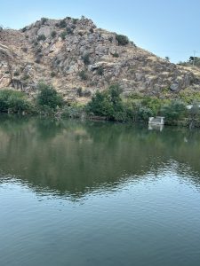 A serene landscape featuring a calm body of water reflecting the surrounding scenery. On one side, there's a rocky hillside adorned with patches of dry grasses and sparse vegetation. Clusters of green trees line the water's edge, while the smooth surface of the water mirrors the hill and trees, creating a tranquil atmosphere.