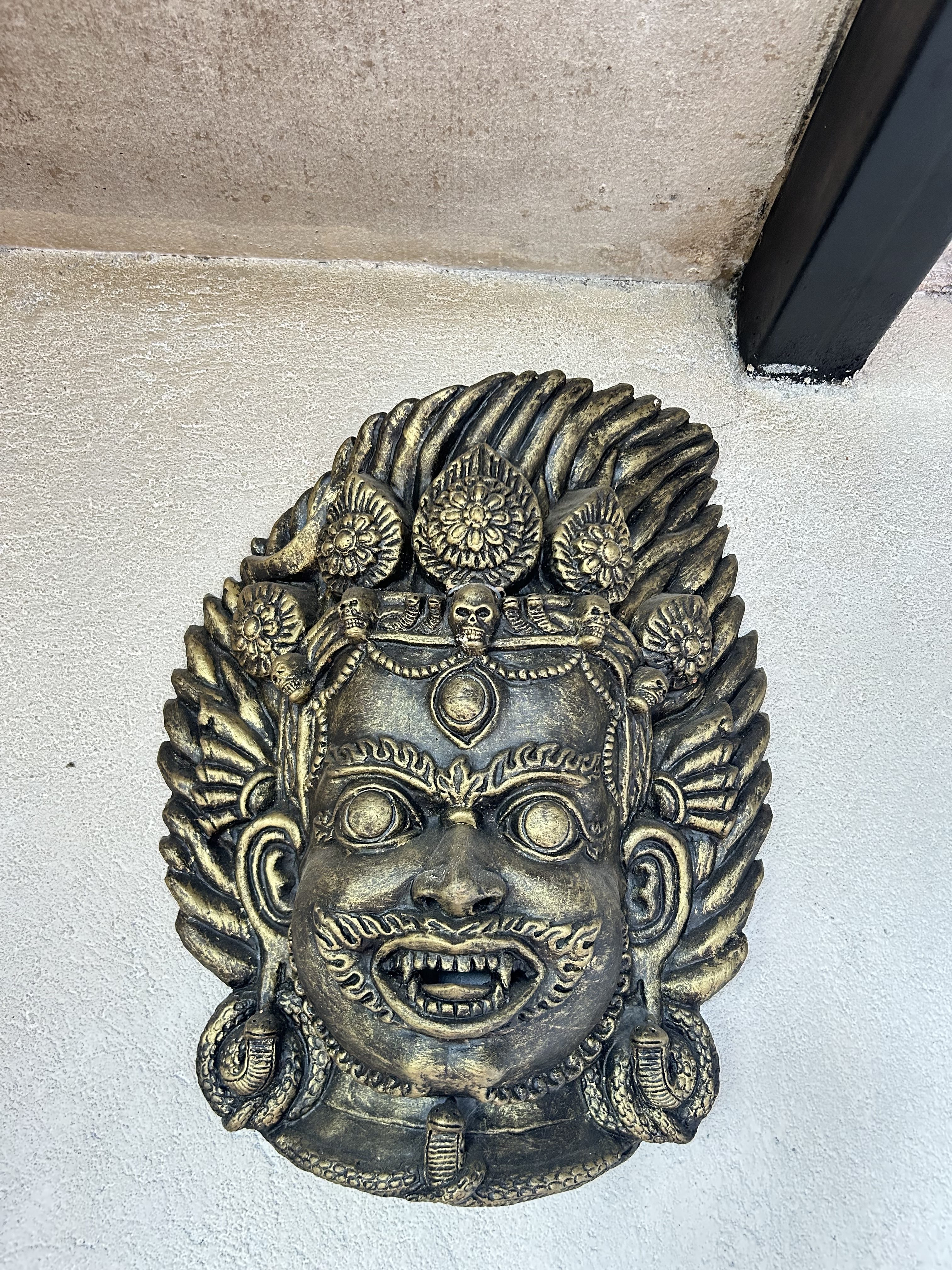 Ornate bronze mask on a wall depicts a fierce, mythological face with wide eyes and bared teeth, adorned with intricate patterns and ornamental details.