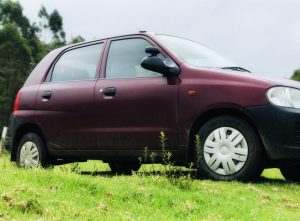 
A maroon compact car is parked on a grassy field, partially on a dirt patch. 