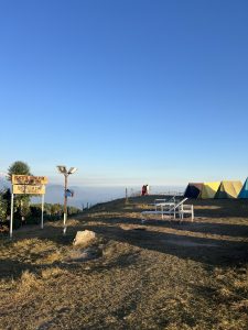 Tents on a hillside under a clear blue sky, with a "Peaceful Himalayan Zone" sign. Two people stand near the edge, overlooking distant mountains.
