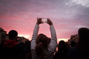 Crowd of people watching a sunset in the distance with one person holding their arms outstretched to take a photo. 