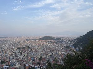 Panoramic view of a sprawling cityscape under a vast sky, with densely packed buildings, a central green hill, and distant mountains.