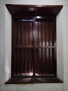 A traditional wooden window with thick shutters allows a narrow beam of light to enter the room. The simple design and aged wood reflect the architectural style of the historic Arakkal residence. Captured at Arakkal Museum, Arakkalkettu, Kannur, Kerala.