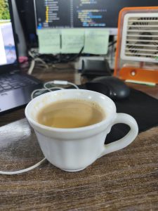 

A white cup of tea sits on a wooden desk, surrounded by a laptop, sticky notes, and a small fan.