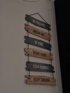 Wall hanging with inspirational messages on wooden slats: "Be Positive," "Dream Big," "Be Kind," "Work Hard," "Stay Humble," "Keep Smiling." Calm and motivational ton