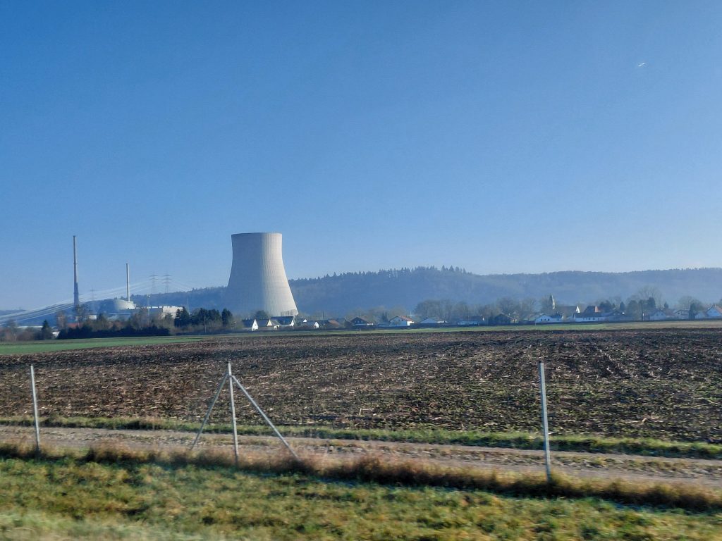 A wide landscape featuring a clear blue sky and distant hills. In the foreground, there’s a flat field with recently turned soil. To the left, a gravel road runs alongside a fence, leading toward a cluster of small, white houses. In the background, a large cooling tower, taller than any nearby structures, stands prominently. – Kernkraftwerk Isar 2