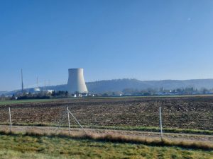 A wide landscape featuring a clear blue sky and distant hills. In the foreground, there’s a flat field with recently turned soil. To the left, a gravel road runs alongside a fence, leading toward a cluster of small, white houses. In the background, a large cooling tower, taller than any nearby structures, stands prominently. – Kernkraftwerk Isar 2
