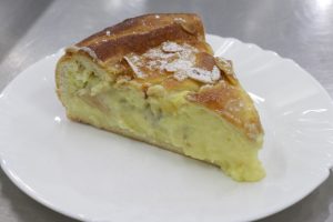 Slice of creamy custard pie with a golden crust, topped with powdered sugar and almond flakes on a white plate. 
