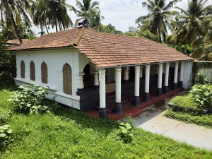 An aerial view showcases a traditional white building with a classic red terracotta-tiled roof, surrounded by lush green lawns, tropical plants, and swaying coconut palms. The long veranda, lined with elegant white pillars, reflects the heritage architecture of the historic Arakkal Palace Complex. Captured at Ayikkara, Kannur, Kerala.