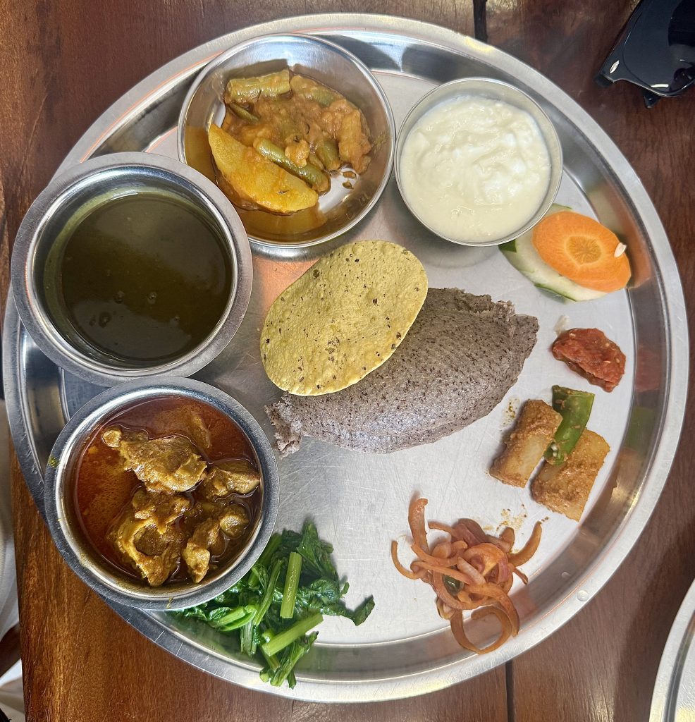 A traditional Nepali thali on a metal plate featuring steamed millet bread, curry, spinach, lentil soup, yogurt, pickles, and carrot slices.
