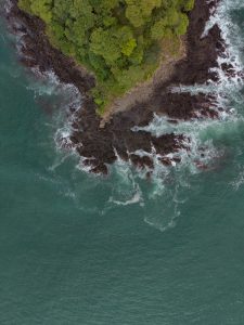 An aerial view of a rocky coastline covered with dense green trees, surrounded by turquoise ocean water. White waves crash against the dark rocks, creating foam patterns along the shore.