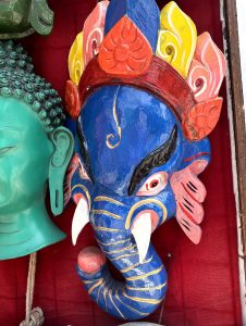 Hand-carved blue wooden god Ganesh mask with colorful floral crown at a Kathmandu market stall. 