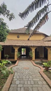 
A rustic house with a thatched roof and yellow walls, framed by lush greenery and potted plants, exudes a serene, tropical vibe on a cloudy day.