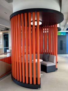 Cozy contemporary office nook with a circular white table and grey sofa, framed by stylish vertical orange wooden slats.