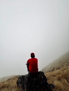 Lone trekker in a red jacket sitting on a rock gazing into a misty Himalayan hillside 