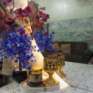 Decorative indoor display featuring vibrant blue and pink artificial flowers in glass vases, a gold ornamental candle holder, and a softly lit mirror on a patterned tabletop, creating a warm and elegant home decor scene.
