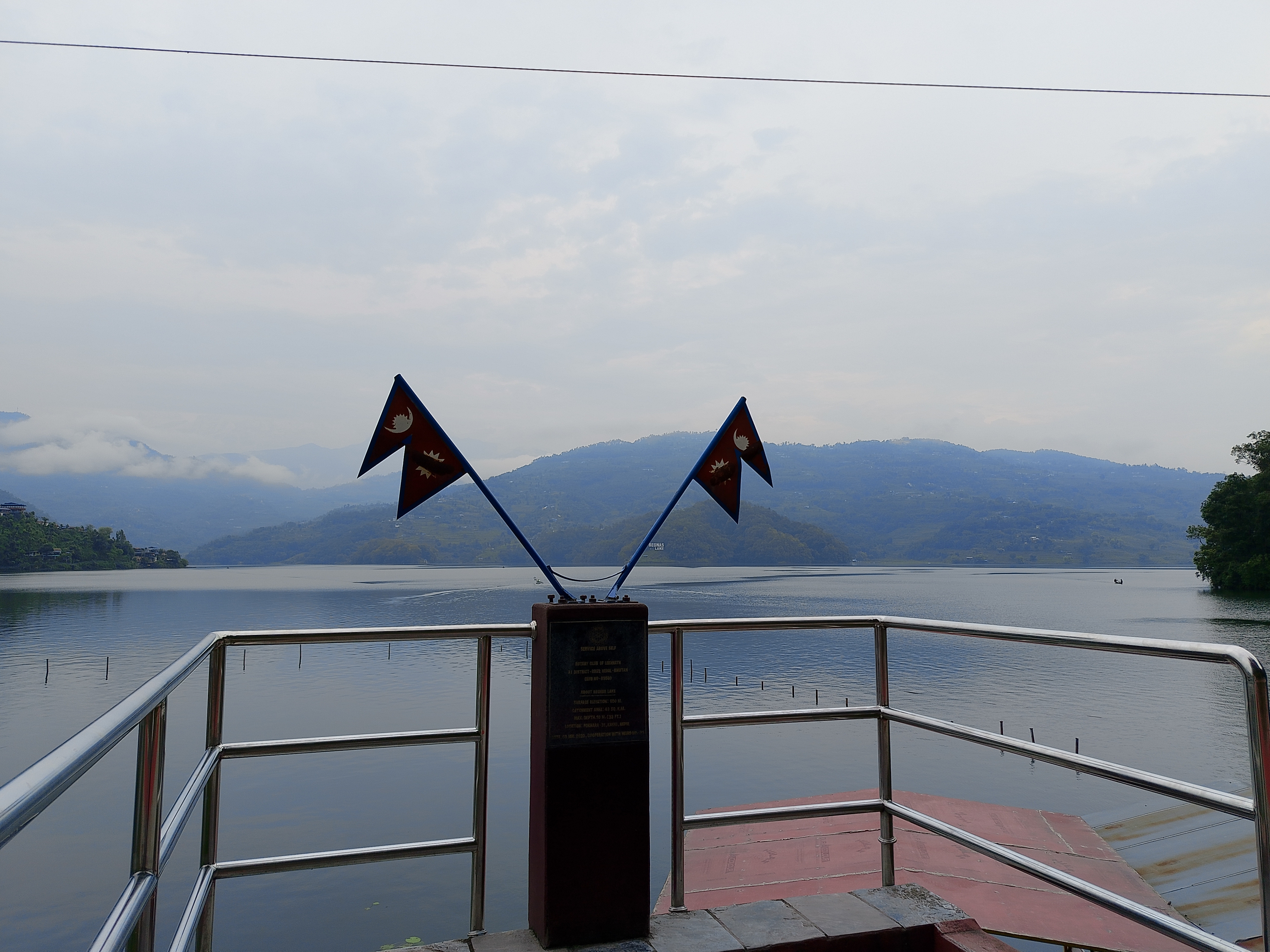 View from the bow of a boat on Begnas Lake, featuring two crossed Nepali flags mounted on a memorial plaque, with calm blue water, forested hills, and low clouds in the background, Pokhara, Nepal.