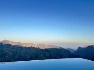Infinity pool edge overlooking forested hills and a hazy valley under a clear blue morning sky.
