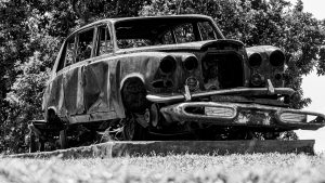 Black and white fine art photograph capturing the historic, rusted wreckage of the Kabaka's classic Rolls-Royce, displayed on a concrete plinth at Mengo Palace with dense trees in the background.
