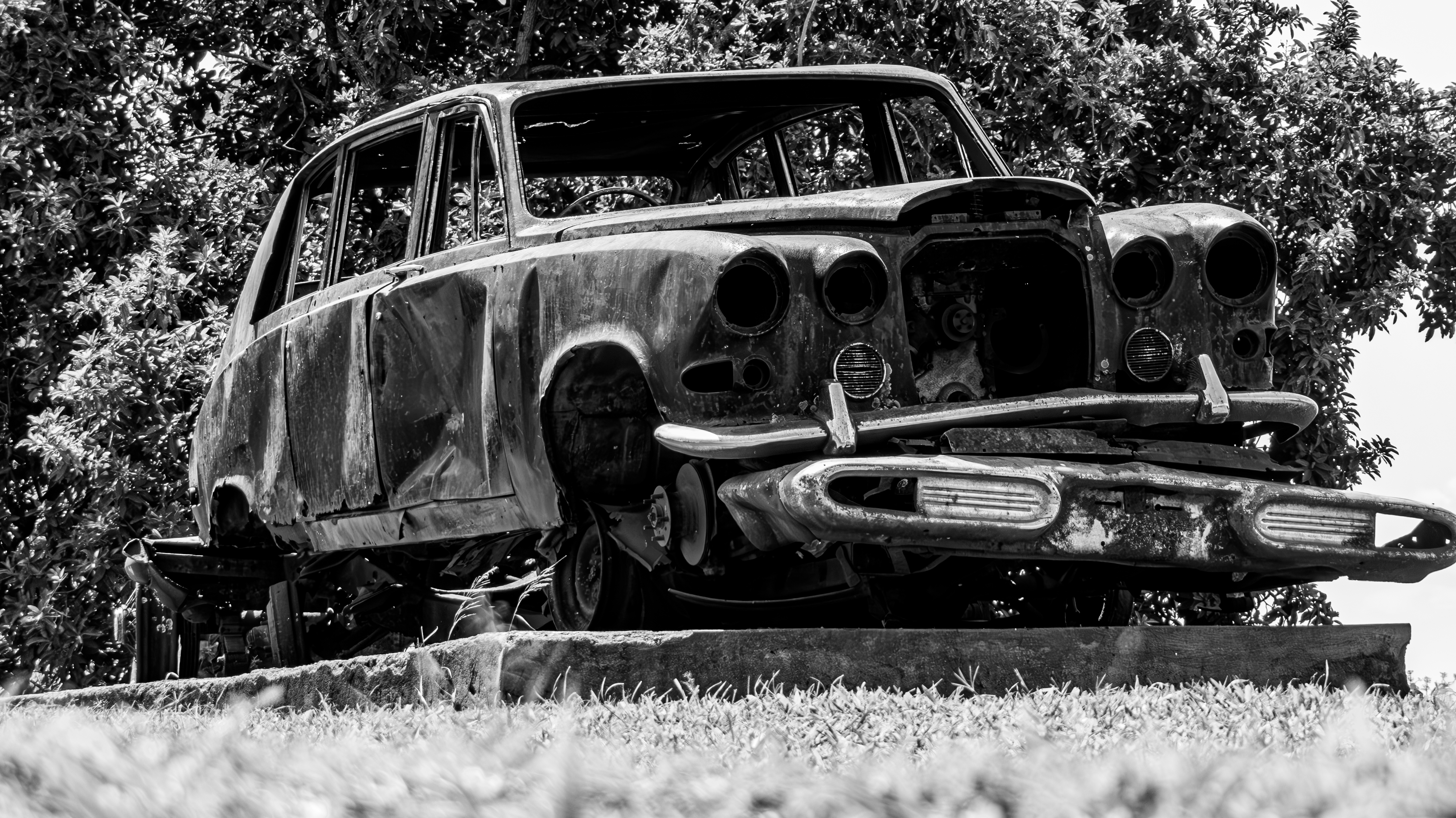Black and white fine art photograph capturing the historic, rusted wreckage of the Kabaka's classic Rolls-Royce, displayed on a concrete plinth at Mengo Palace with dense trees in the background.