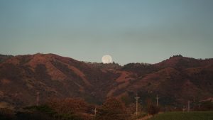 Landscape of mountains at sunset, with warm tones and a full moon visible between two hills. Trees, power poles, and vegetation appear in the foreground, while a clear sky fills much of the image.