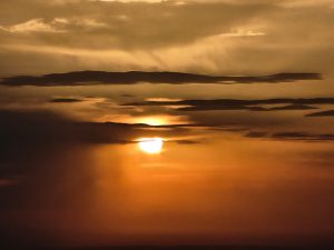 Sunset with a warm orange and yellow sky, scattered dark clouds partially obscure the sun.