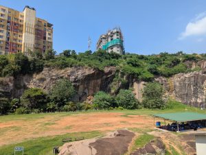 Sunny urban landscape with a rocky grassy foreground, colorful buildings, a construction site, and people gathered under a covered area beneath a clear blue sky.
