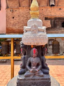 A small stone chaitya (Buddhist stupa) with a golden gilded top supported by a multi-armed deity figure base with red vermillion offerings, surrounded by prayer wheels, Nepal. 