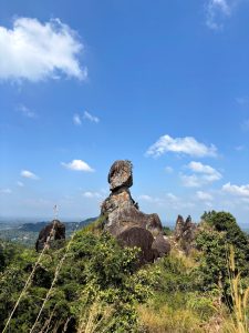 Distinctive rock formation rising above greenery and hills beneath a blue sky. 