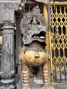A large ornate gilded bronze ritual incense burner or water vessel (kalash) topped with a fierce makara head and deity figure, flanked by carved stone columns at a Newari temple, Nepal. 