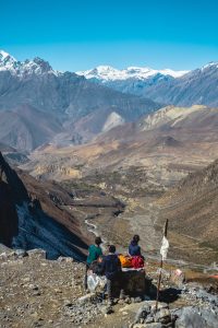 Three trekkers resting beside a prayer flag on a rocky high-altitude perch, gazing over a vast layered canyon valley toward snow-capped Himalayan peaks