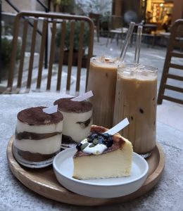 A cozy cafe table with two tiramisu cups, a slice of blueberry cheesecake on a plate, and two iced coffees on a wooden tray. Background shows chairs and greenery.