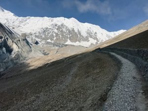 A rugged, winding mountain trail leads through rocky terrain toward snow-capped peaks, under a clear blue sky, evoking a sense of adventure.