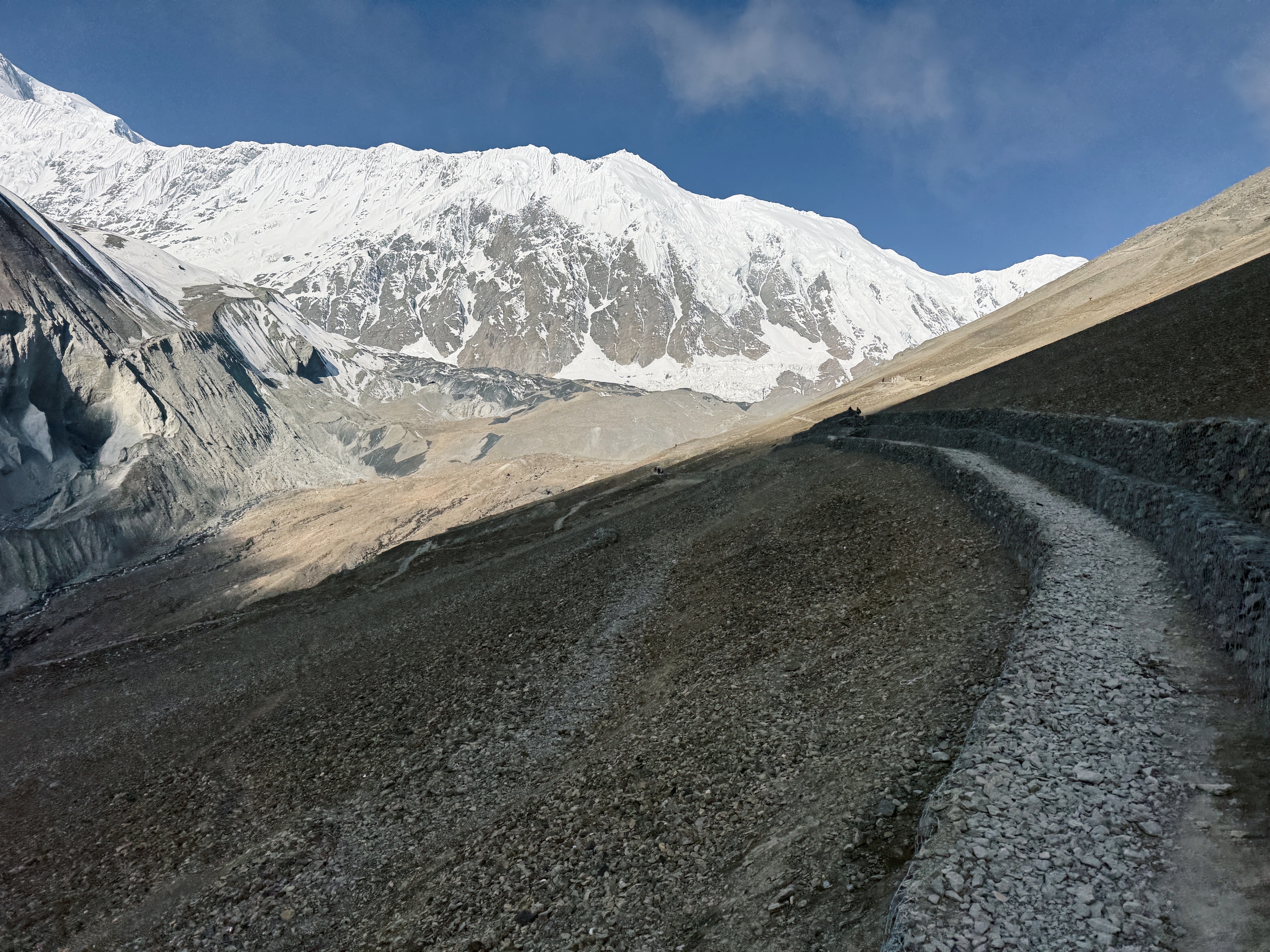 A rugged, winding mountain trail leads through rocky terrain toward snow-capped peaks, under a clear blue sky, evoking a sense of adventure.