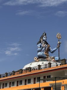 A large blue statue of Lord Shiva with a trident sits atop a building under a clear blue sky. The figure exudes calmness and power, surrounded by architectural details. 

