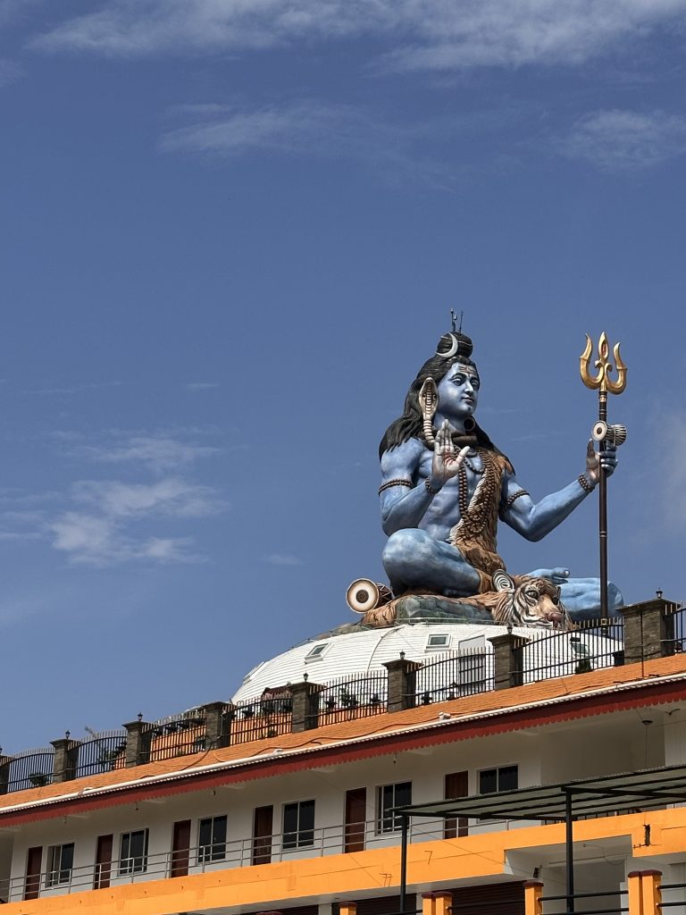 A large blue statue of Lord Shiva with a trident sits atop a building under a clear blue sky. The figure exudes calmness and power, surrounded by architectural details.