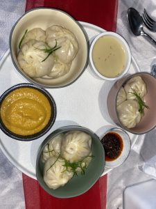 A round white plate is set against a backdrop with red and gray colors. On the plate, there are three small bowls filled with dumplings, each one featuring a delicate pleated design. One bowl is light green and contains three dumplings topped with thin, fresh green herbs. 
