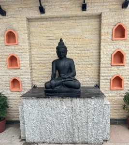 A serene black stone Buddha statue in a meditative pose on a pedestal, set against a beige brick wall with orange alcoves, surrounded by potted plants.
