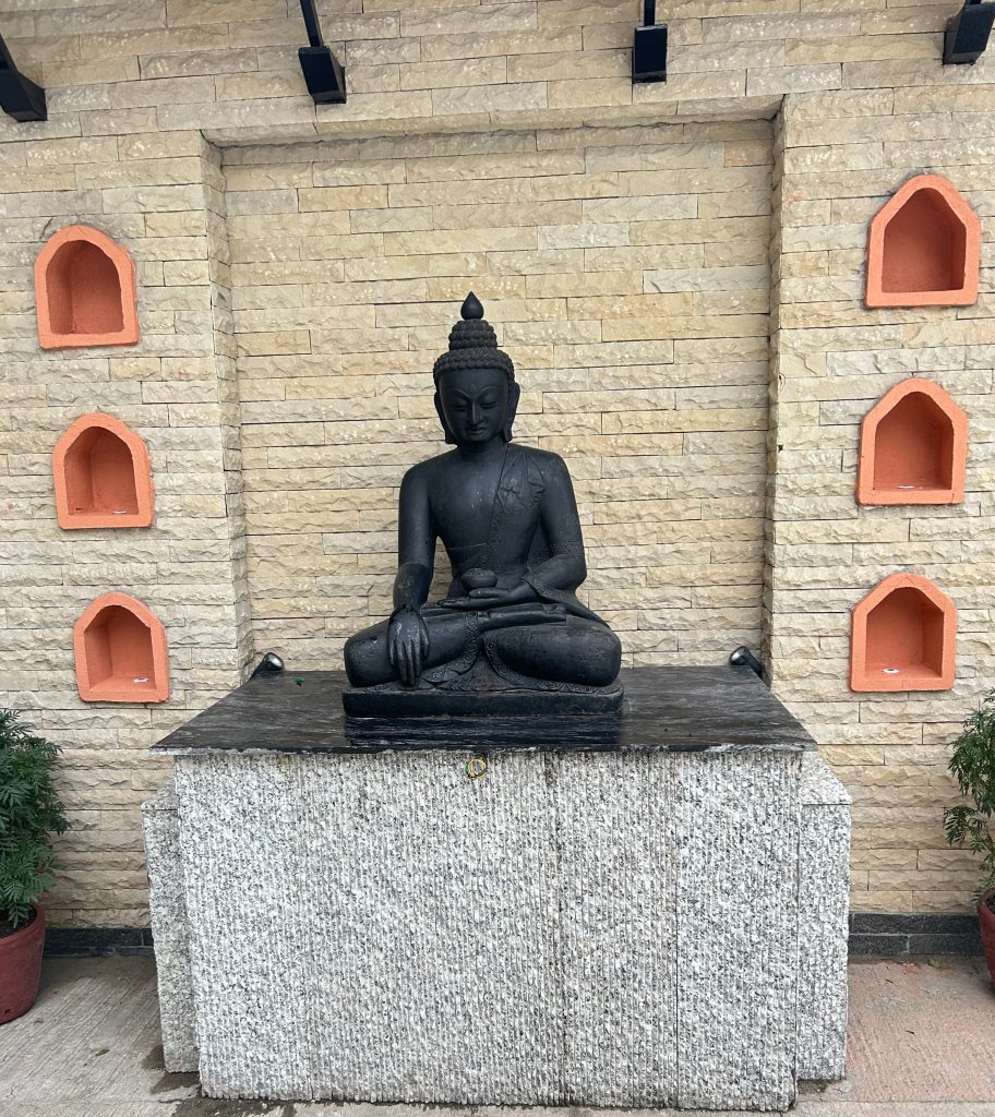 A serene black stone Buddha statue in a meditative pose on a pedestal, set against a beige brick wall with orange alcoves, surrounded by potted plants.