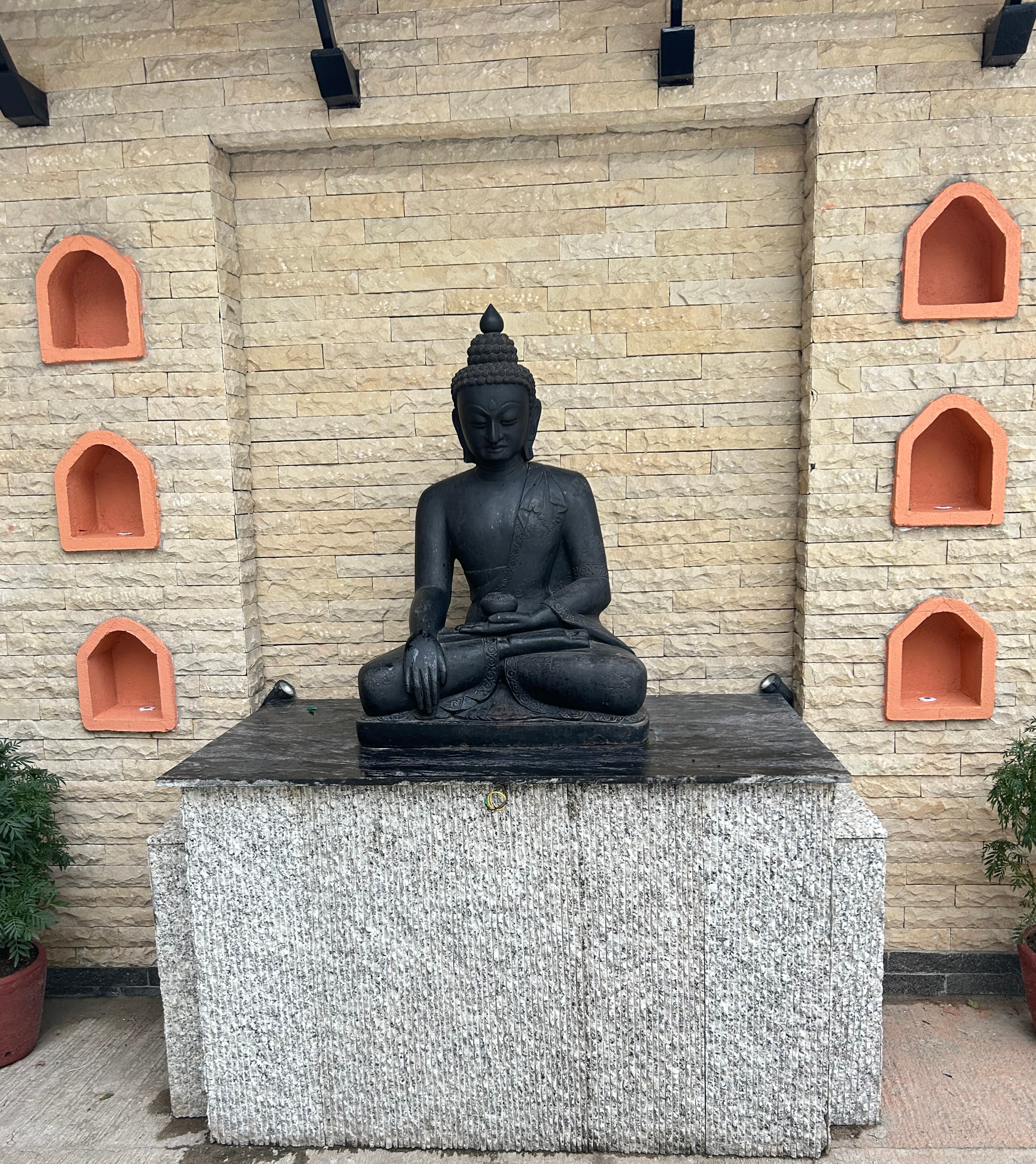 A serene black stone Buddha statue in a meditative pose on a pedestal, set against a beige brick wall with orange alcoves, surrounded by potted plants.