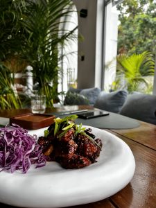 A dish of glistening glazed meat garnished with green onions, paired with shredded purple cabbage, on a white plate in a cozy, plant-filled cafe.