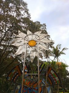 A public monument showcasing a large metal sunflower with a yellow stained-glass center, mounted in front of abstract patterned mosaic glass panels. 
