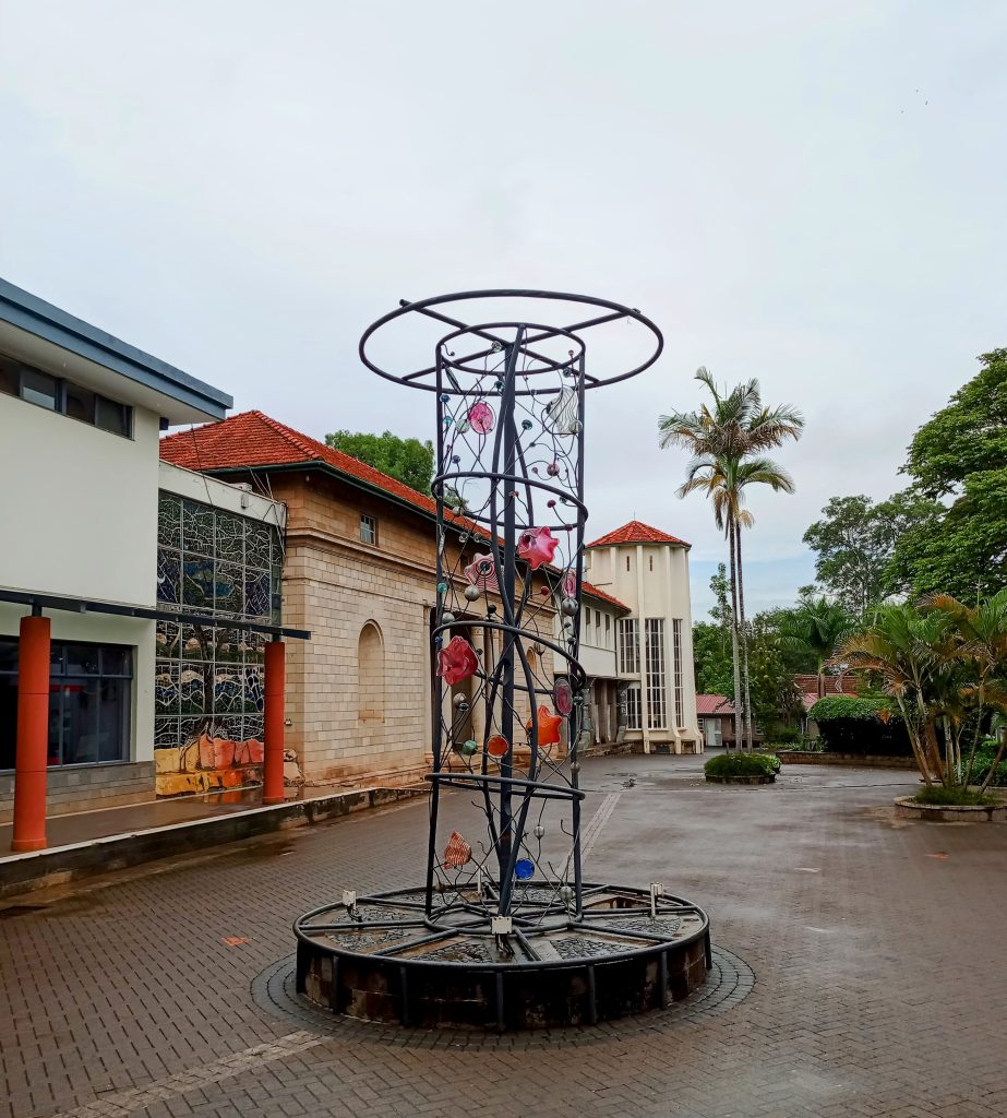 A tall outdoor metal sculpture featuring a spiral cage design accented with colorful glass ornaments, situated in a paved courtyard.